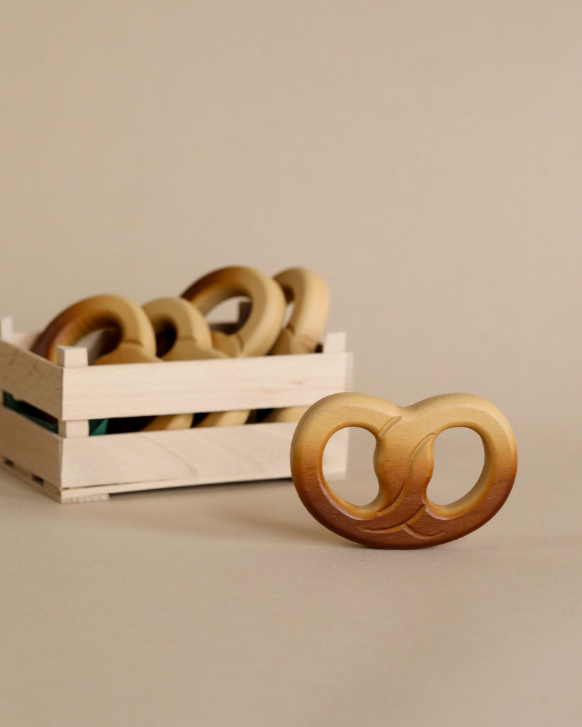 A Erzi Pretzel Pretend Food teether in the foreground with more handcrafted toys in a white crate against a beige background, focusing on sustainable baby products.