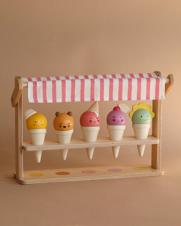 A toy Ice Cream Scoops and Smiles stand displaying five colorful solid wood ice cream cones with cute, smiling faces on the scoops, arranged on a two-tiered shelf with a striped awning above.