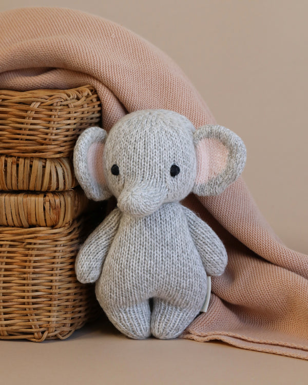 A Cuddle + Kind Baby Elephant made from heirloom-quality Peruvian cotton yarn, with large ears and black button eyes, is propped against a stack of woven wicker baskets. It is partially covered by a soft pink blanket hanging to the left. The background is a warm beige color.