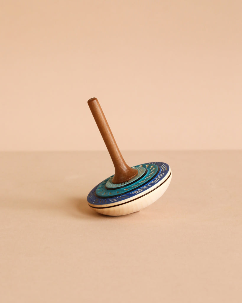 The Mader Bonbon Spinning Top - Blue features a decorated blue and white circular base, an embossed pattern, and a brown wooden handle, shown upright on a beige surface with a matching beige background.