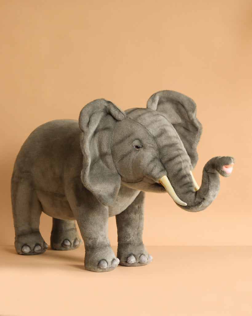 The Ride-On Elephant is a large plush toy with realistic features, artisan hand-sewn in soft gray fabric, featuring white tusks and a raised trunk, standing on a plain light brown background.
