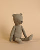 A brown, fabric Maileg Teddy Dad with a vintage look and jointed limbs is sitting against a beige background. The bear has minimalist facial features, including small button-like eyes and a stitched nose and mouth. One leg is extended while the other is bent at the knee, showcasing its soft linen texture.
