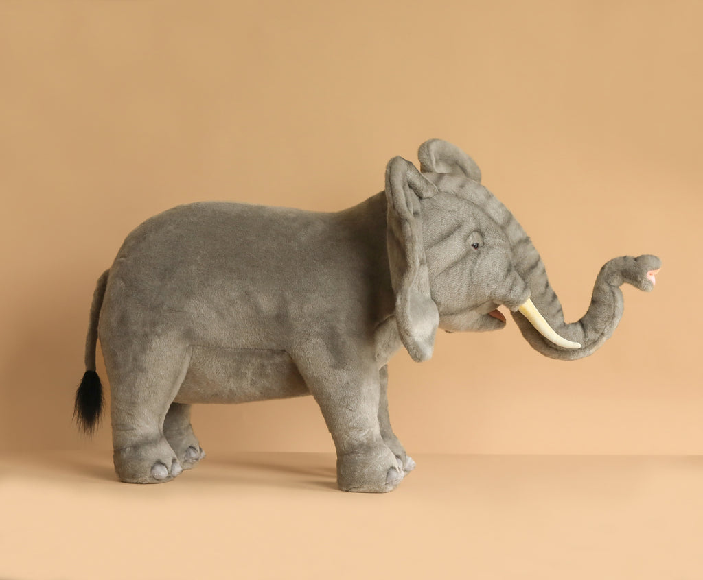 The Ride-On Elephant is a large, artisan hand-sewn plush with realistic features—white tusks, raised trunk, and slightly open mouth—shown standing on a plain light brown background.