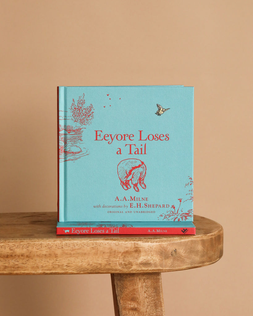 A blue hardcover "Winnie-the-Pooh: Eeyore Loses a Tail" book with red illustrations and text by A.A. Milne stands upright on a wooden stool, with another copy lying flat beneath it against a beige background.