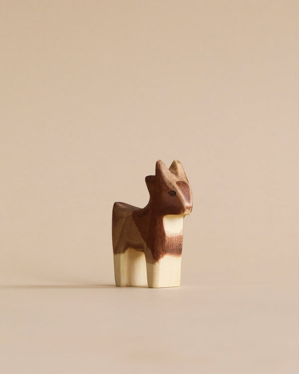 A handmade Holzwald small goat, crafted with different shades of brown, stands on a light beige background. This high-quality toy shows detailed carving, emphasizing the goat's distinct features.