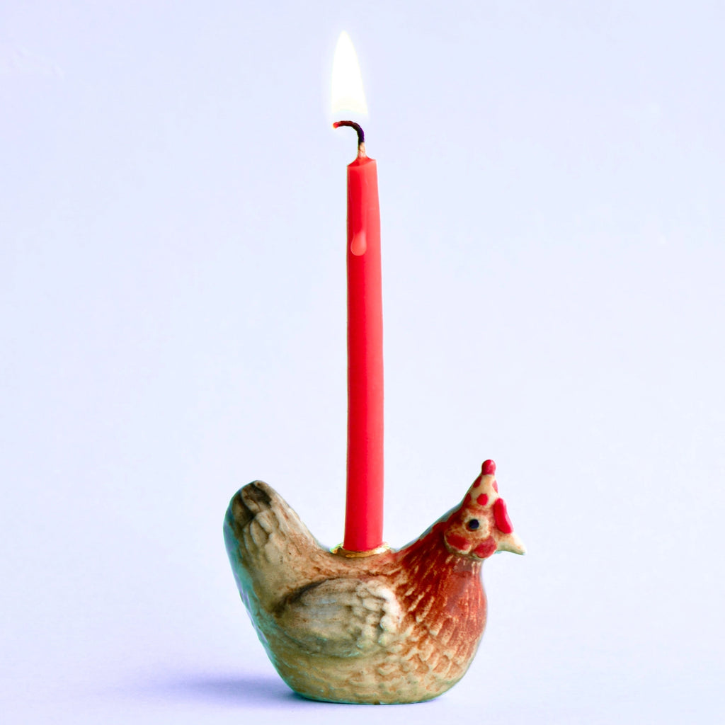 A red candle lit and held upright by a hand-painted porcelain rooster cake topper, against a light blue background.