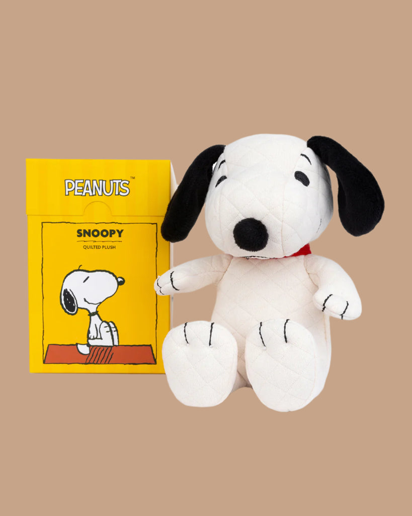 The Peanuts Snoopy Plush Toy, featuring a white body, black ears, and a red collar, is presented in a yellow gift box with a Snoopy illustration on one side, all against a plain brown background.