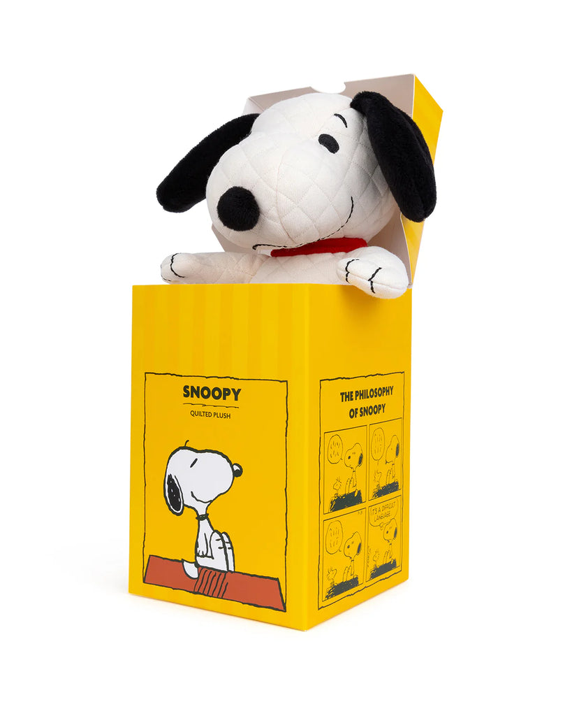 A Peanuts Snoopy Plush Toy peeks out from a yellow box adorned with whimsical illustrations, labeled "Snoopy Quilted Plush" and "The Philosophy of Snoopy," making it a charming addition to any collection.