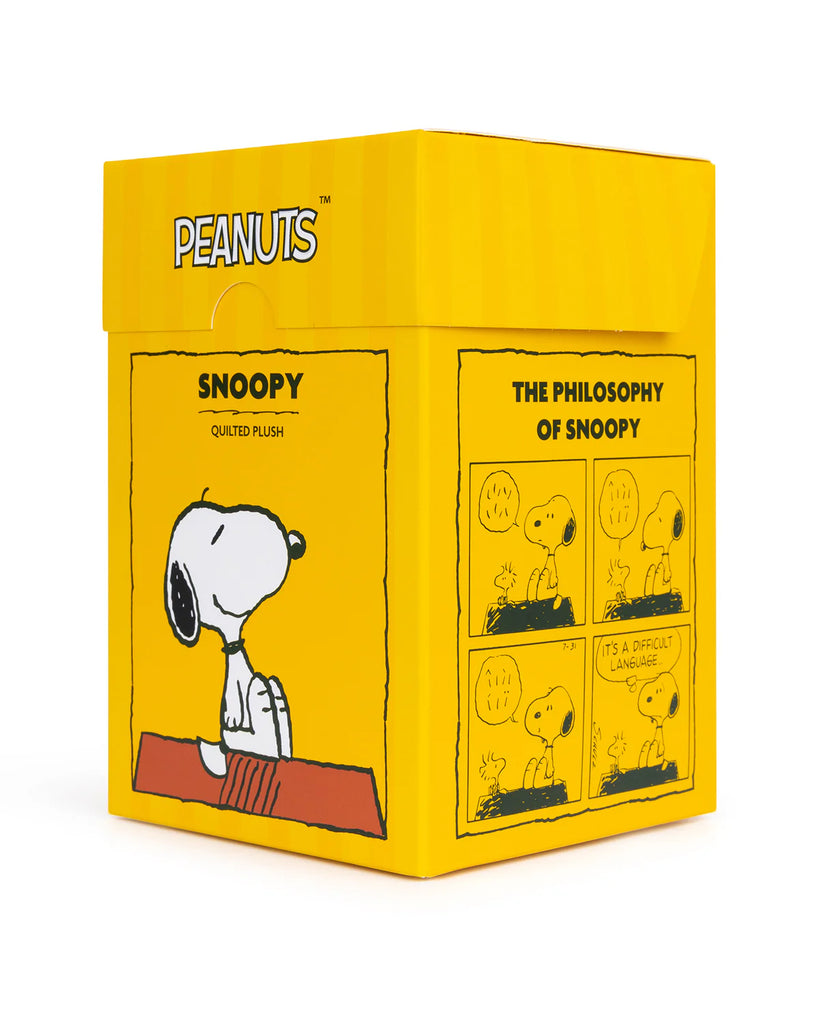 The Peanuts Snoopy Plush Toy In Gift Box features a quilted plush Snoopy in a yellow, Snoopy-branded box. One side displays a comic strip with Snoopy and Woodstock discussing language challenges, adding charm to any plush collection.