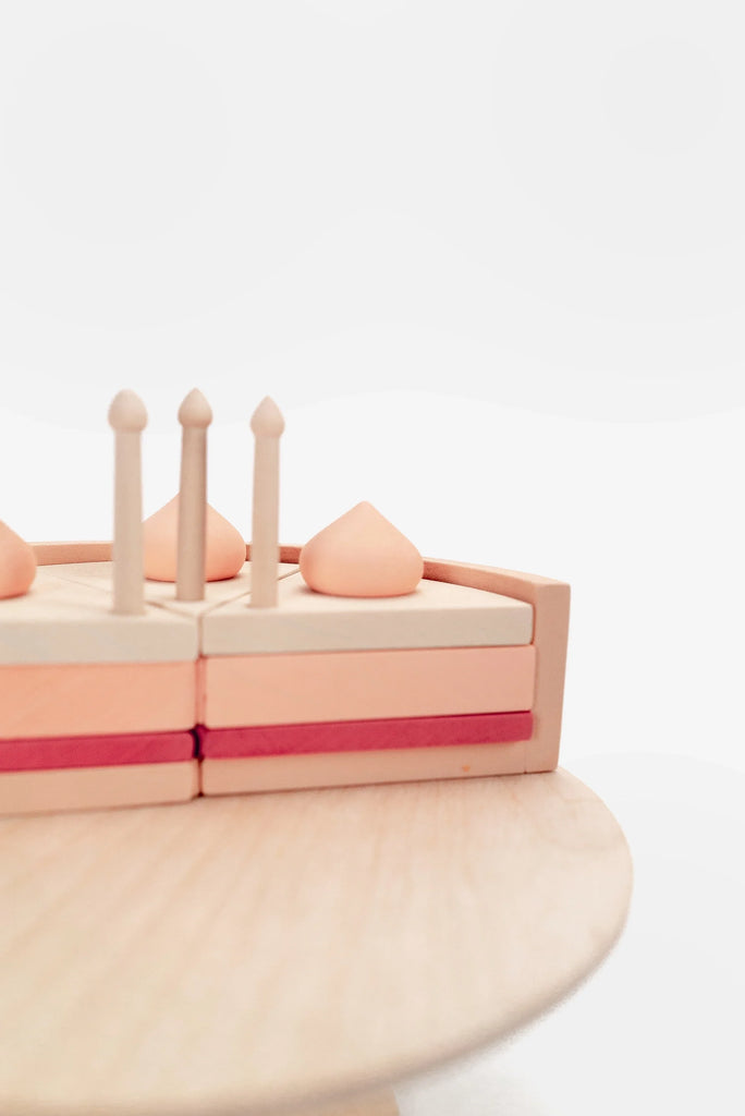 A minimalistic photo featuring a wooden Handmade Strawberry Layer Cake On A Stand toy with various shaped pieces, including cones and a semi-sphere, arranged on a circular base against a white background. This educational toy is painted with