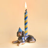 A Dachshund Cake Topper of a black and tan dog wearing a blue polka-dotted party hat, with a lit blue and white striped candle on its back, against a beige background.