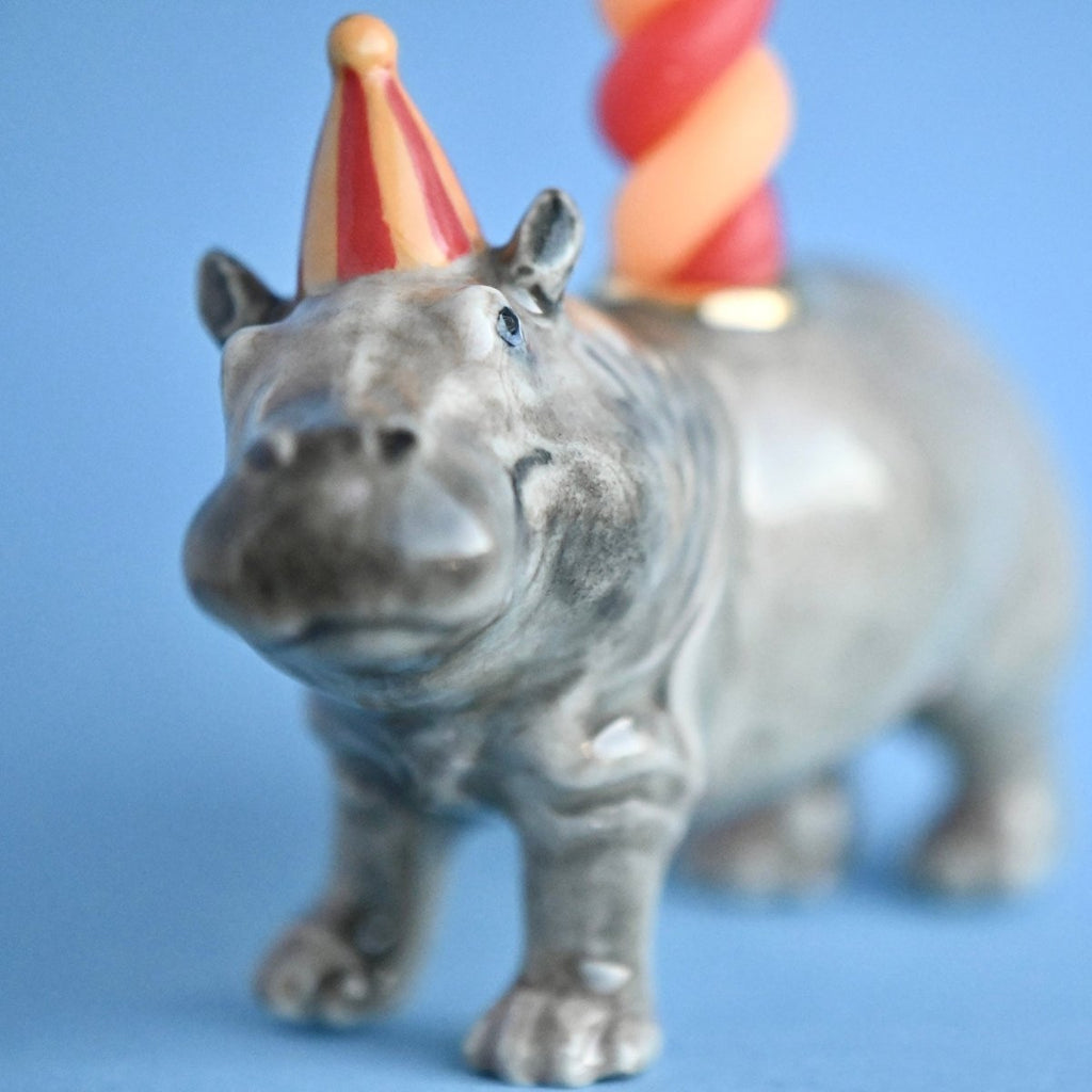 A Hippo Cake Topper, hand painted and adorned with a party hat and a colorful ice cream cone on its back, set against a plain blue background. Focus is on the hippo's face.