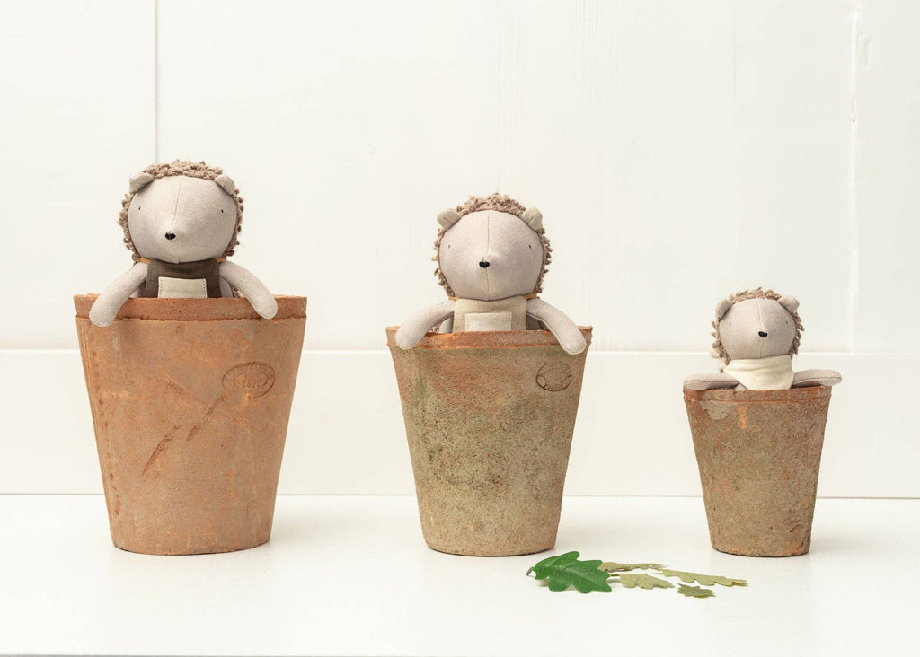 The whimsical tableau features three "Heather The Hedgehog" plush toys in terracotta pots of varying sizes, with two small green felt leaves set against a white background.