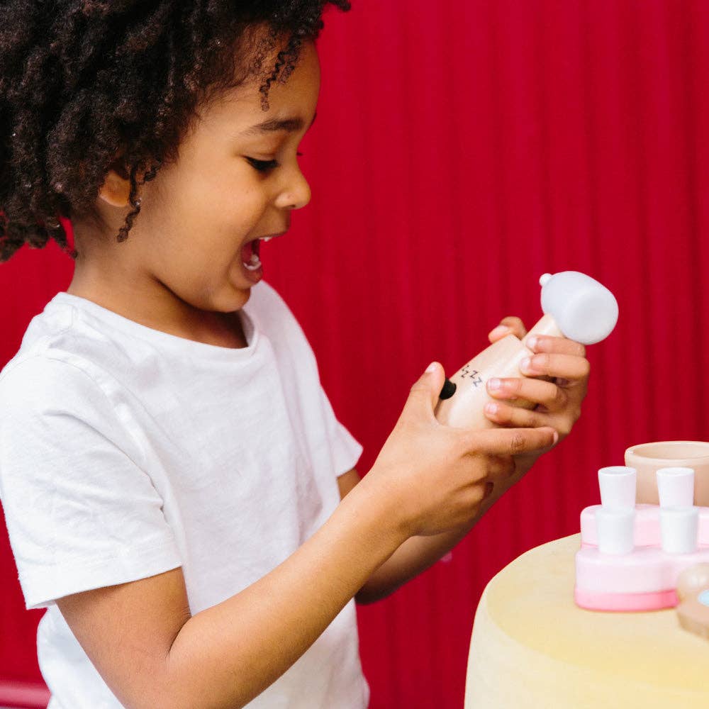 A young child wearing a white T-shirt excitedly holds a wooden toy bottle from the Wooden Dentist Set, examining it with interest. Several small bottles and containers from the set are on a yellow table, set against a bright red background.