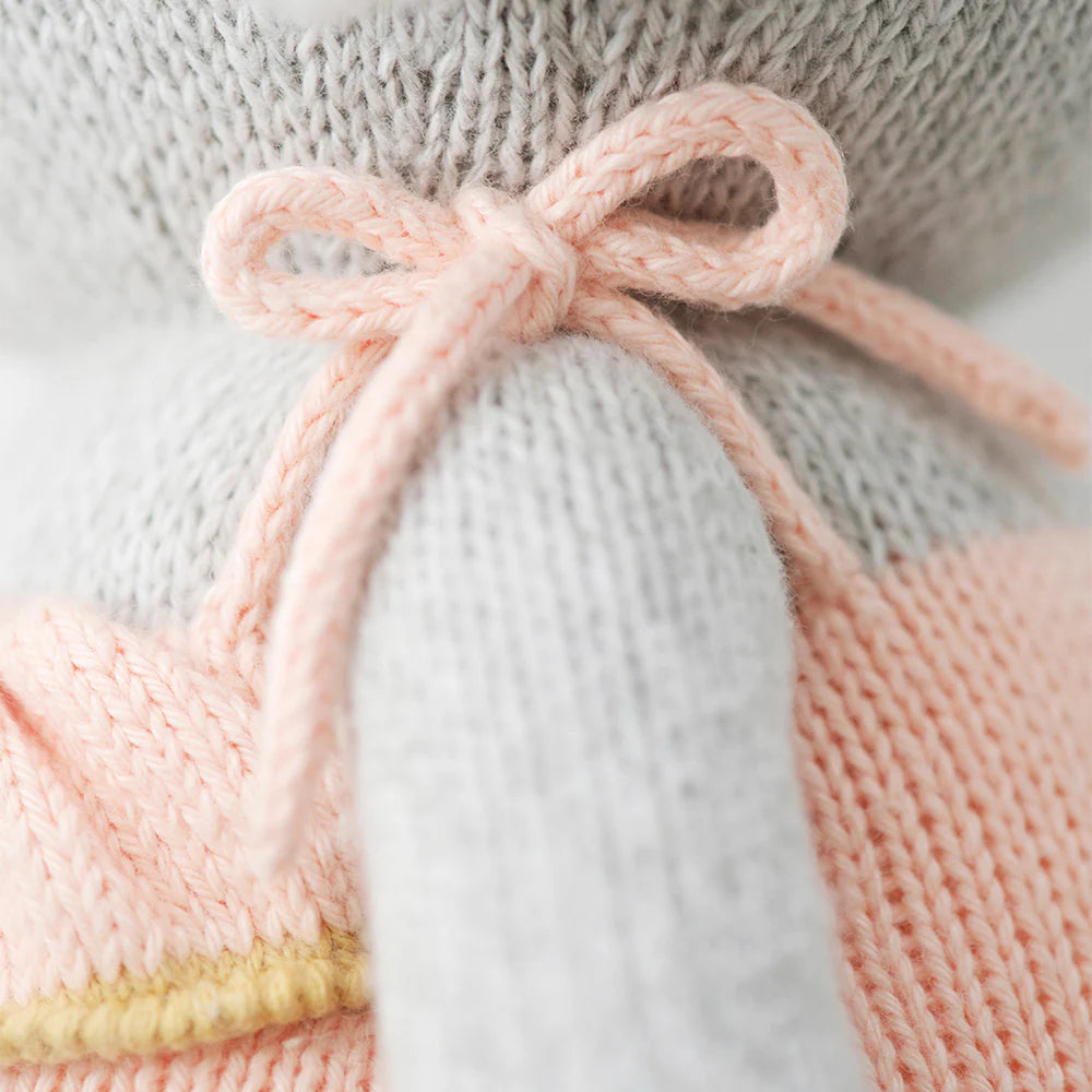 Close-up of Cuddle + Kind Eloise the Elephant, a soft hand-knit stuffed animal in a peach sweater with a pale pink yarn bow. The detailed knit texture is visible. Each doll provides 10 meals to children in need.