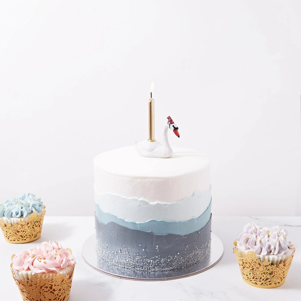 A hand-painted porcelain ombre cake with shades of blue, decorated with a lit candle and a small figure of a diver on top, surrounded by assorted cupcakes on a white background, featuring a Swan Cake Topper.