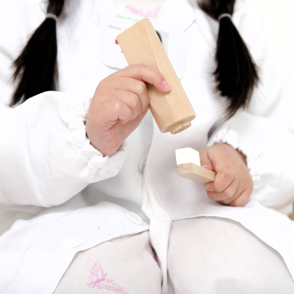 A child in a white coat, playing pretend with the Wooden Dentist Set, holds two wooden toy tools. Her face is out of frame, dark pigtails visible, and her pants display a small butterfly design.