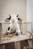 A cuddly Steiff Cow Stuffed Animal with a cheerful face sits on a wooden bench surrounded by hay, inside a cozy room. The cow has black and white patches, distinct horns, and features the iconic Steiff button in its ear.