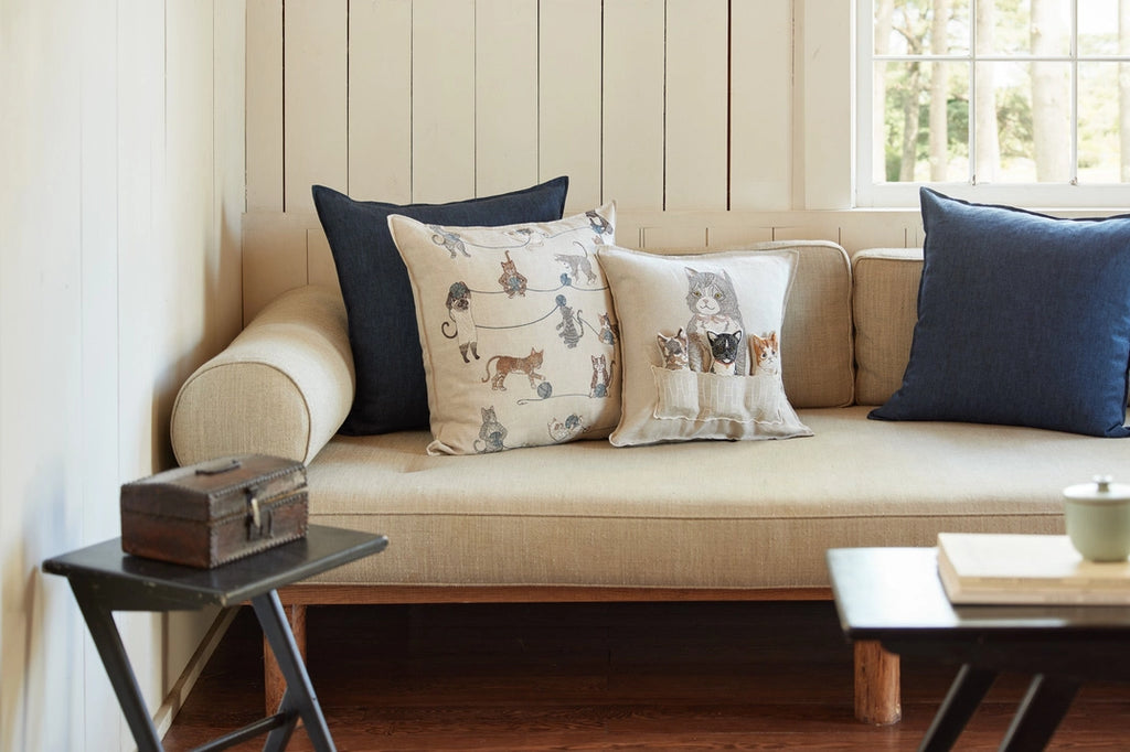 A cozy living room nook featuring a beige sofa lined with various pillows, including one Coral & Tusk Basket of Kittens Pocket Pillow, beside a window and a small black side table with a vintage box.
