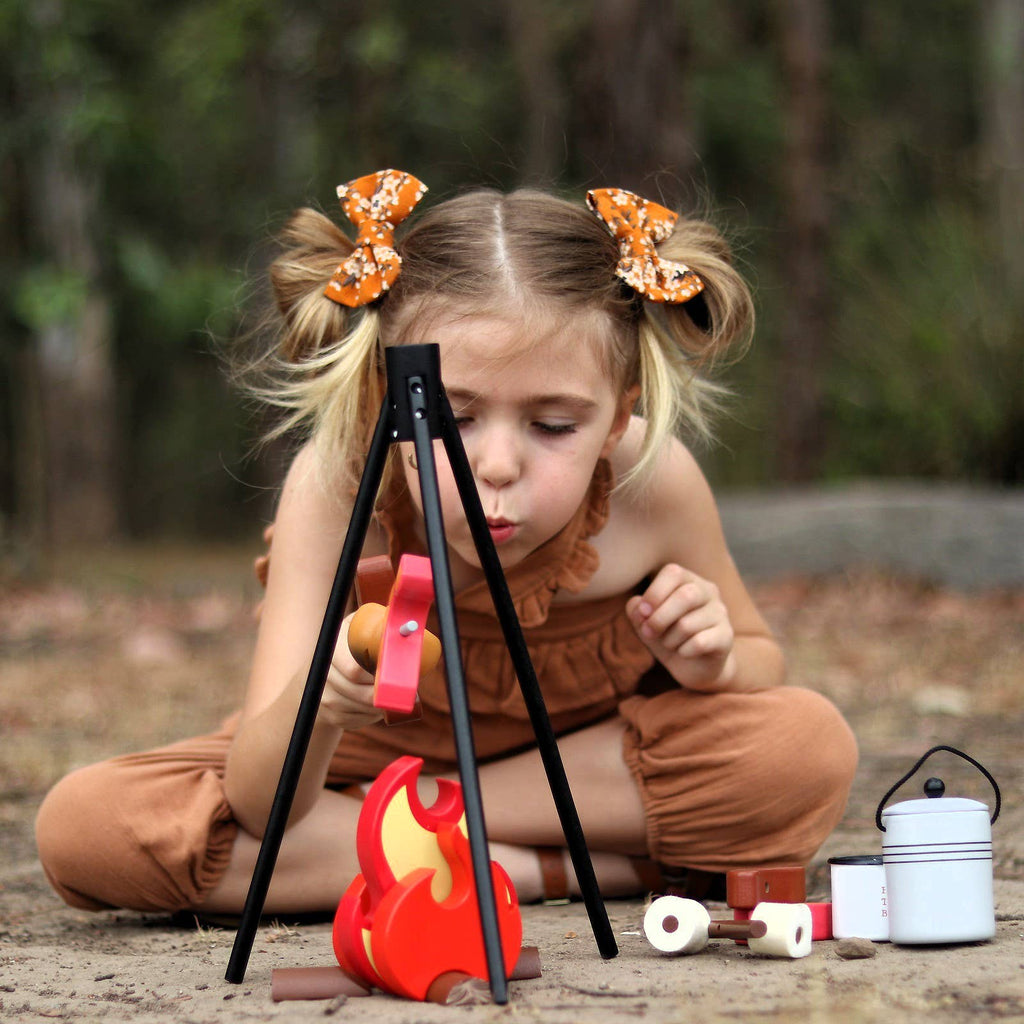 A young child with orange bows enjoys pretend play outdoors, blowing on the Wooden Camp Fire Set, which features a tripod, toy food items, and a small kettle arranged on the ground.
