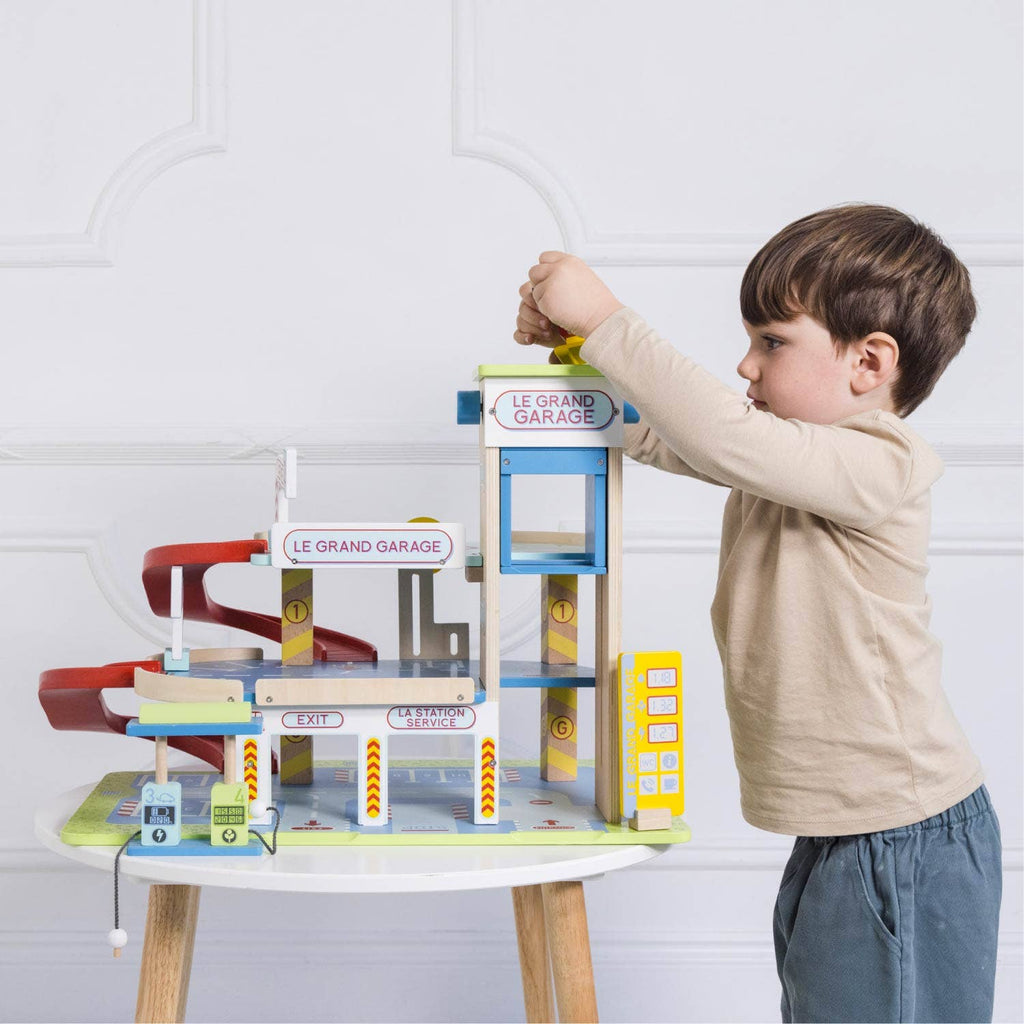 A young boy plays imaginatively with the Le Grand Wooden Garage, placing a car on the top level. This educational toy features ramps, signs, and multiple levels, set on a small table against a white wall.