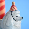 A close-up image of a whimsical white fine porcelain Poodle Cake Topper sporting a striped party hat against a vibrant blue background.