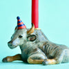 A Ox Cake Topper shaped like a seated bull with a party hat, featuring a beeswax birthday candle inserted on its back against a light blue background.