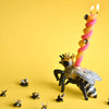 A whimsical Queen Bee Cake Topper wearing a golden crown and a pink spiral candle on its back against a bright yellow background, surrounded by smaller fly figurines.