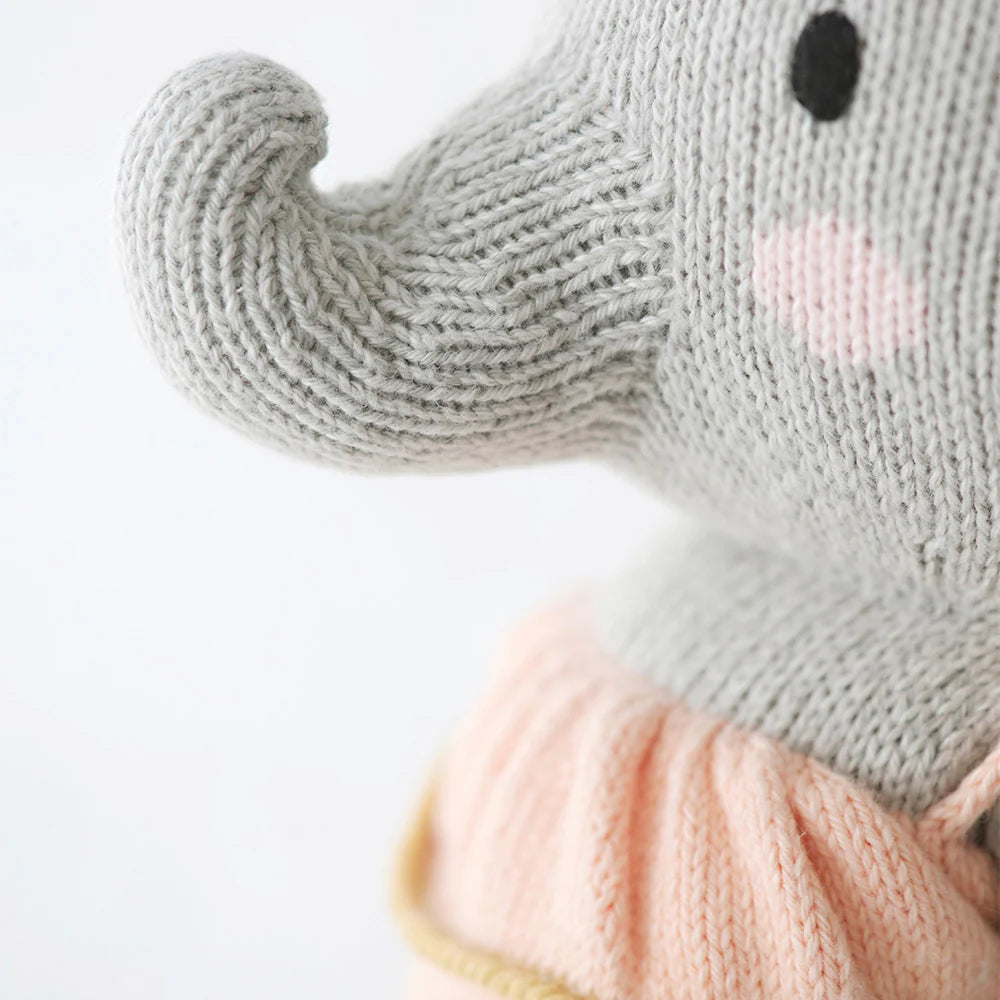 Close-up of Cuddle + Kind Eloise the Elephant, a gray hand-knit doll with pink cheeks in pink and yellow attire. Detailed yarn texture is visible. Every savanna stuffed animal like this provides 10 meals to children in need.