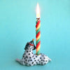 A colorful twisted candle lit on top of a hand painted porcelain Dalmatian Cake Topper against a light blue background.