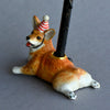 A Corgi Cake Topper figurine of a merry corgi dog wearing a red-striped party hat, lying down with a black walking stick resting atop. The dog has a realistic brown and white coat, hand painted porcelain