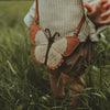 A toddler from behind holding a Donsje Toto Purse in a grassy field. The purse, featuring premium cream metallic suede with orange, black, and white colors resembling butterfly wings, includes an adjustable leather strap.