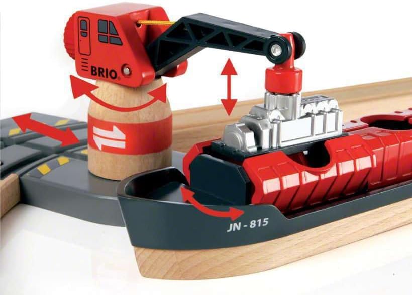 A BRIO crane lifts a silver cargo from a red and black ship labeled "JN-815" in the BRIO World Train Set Cargo Harbour Set, with red arrows showing crane and cargo movement—ideal for imaginative train play.