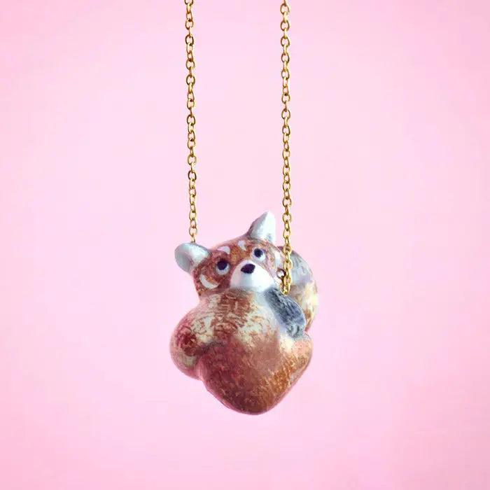 A necklace with a pendant shaped like a small, curled-up fox sculpted in a realistic style. The 24k gold plated fox pendant hangs from a delicate gold chain, showcasing heirloom quality against a solid soft pink background.
Red Panda Necklace:
A necklace with a pendant shaped like a small, curled-up red panda sculpted in a realistic style. The 24k gold plated red panda pendant hangs from a delicate gold chain, showcasing heirloom quality against a solid soft pink background.
