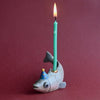 A whimsical candle holder shaped like a Fish Cake Topper, with a lit green birthday candle inserted in the holder, set against a deep red background.