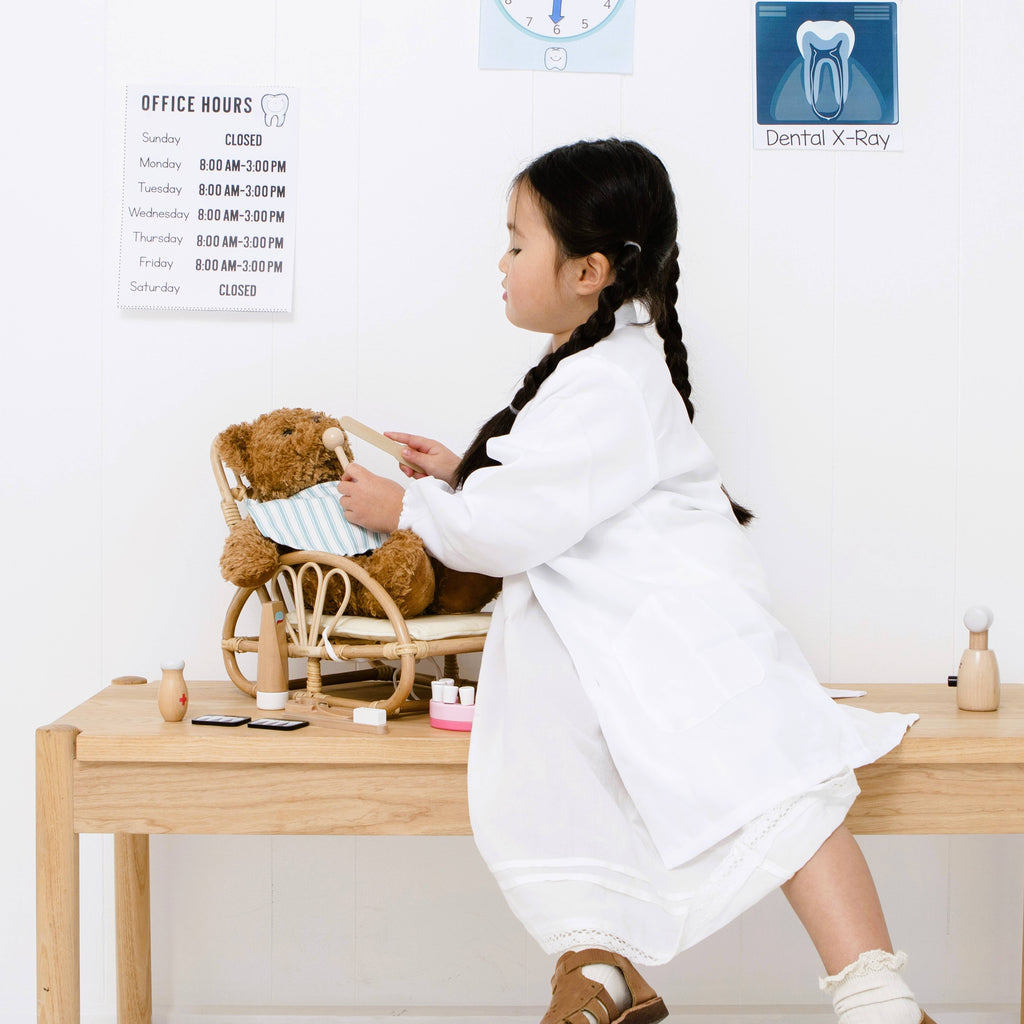 A young girl uses the Wooden Dentist Set to examine a teddy bear in a toy wheelchair, holding a dental tool. Behind her, office hours and a dental X-ray sign are visible on the wall.