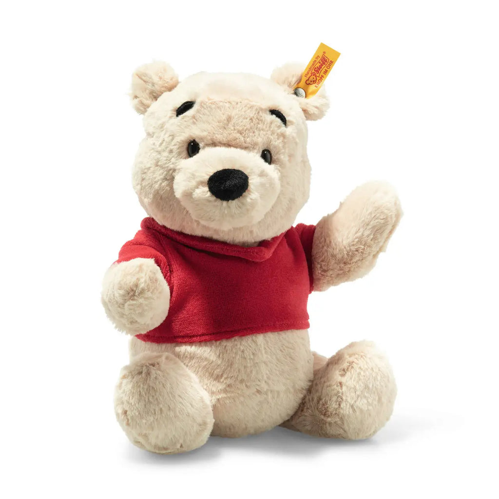 A Steiff plush toy bear with cream-colored fur, wearing a red shirt, sitting on a white background. The bear appears soft and cuddly, with a friendly expression and distinctively features a "Button in".
