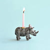 A small Rhino Cake Topper with a pink birthday candle secured on its back, lit and burning brightly, against a soft blue background.