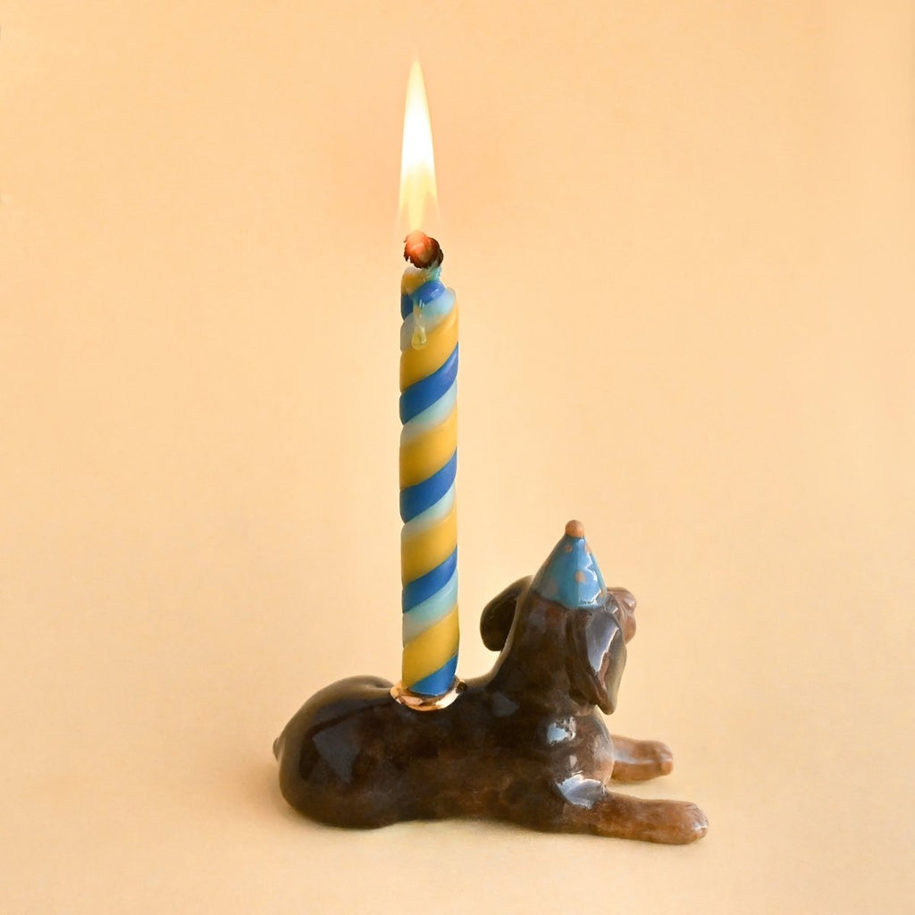 A whimsical hand-painted Dachshund Cake Topper, featuring a blue and white striped candle lit on top, set against a soft peach background.