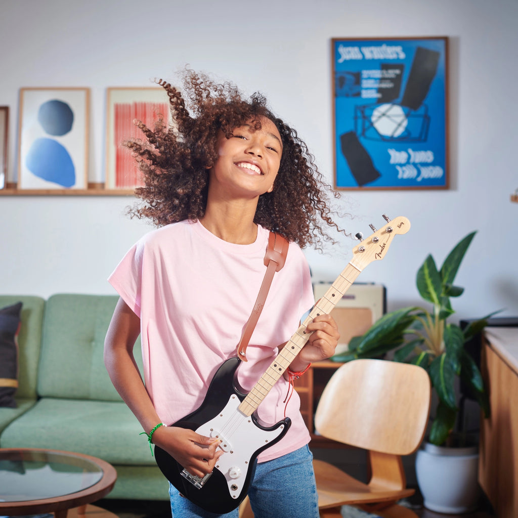 A joyful young woman with curly hair plays a Fender X Loog Stratocaster Electric Guitar indoors. She wears a pink t-shirt and smiles with closed eyes, surrounded by vibrant home decor and plants.