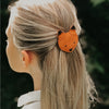 A woman with blonde hair tied back in a ponytail, secured with a Donsje Leather Hair Tie - Fox that's handmade and fairtrade. The setting appears to be outdoors with a lush green background.