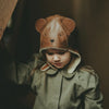 A young child wearing a Donsje Leather Classic Hat - Bear with ears and button eyes, dressed in a green coat with sheep wool lining, stands in a shadowy area, looking slightly downward with a thoughtful expression.
