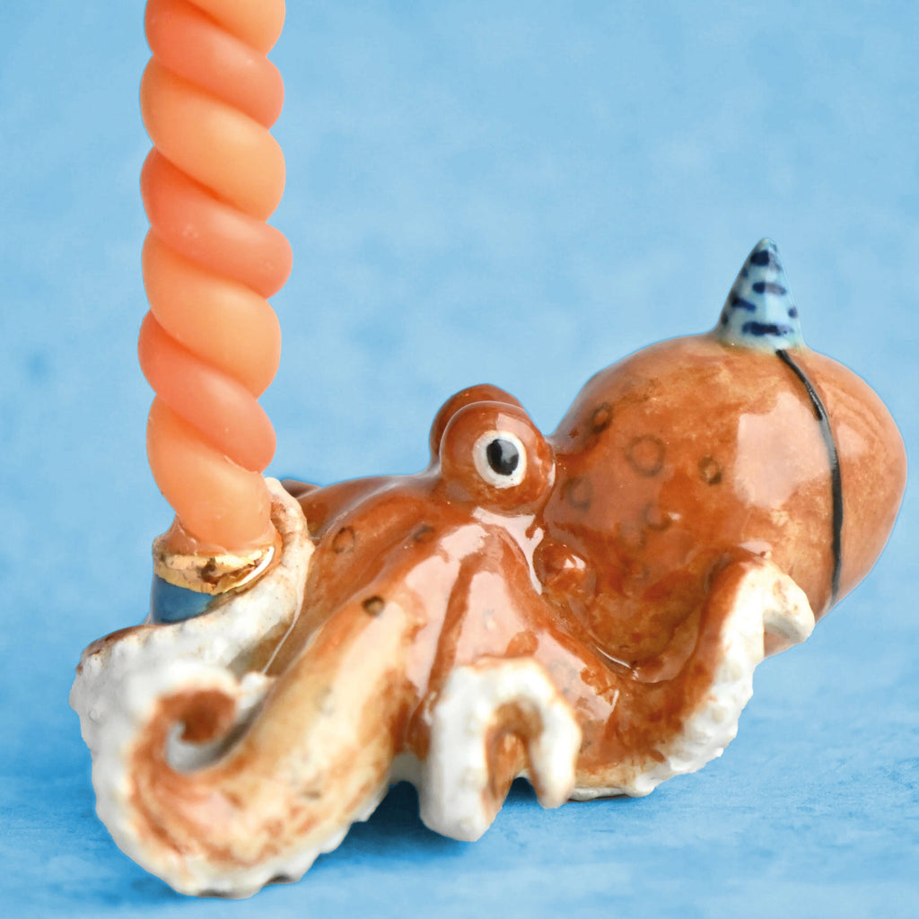 A fine porcelain Octopus Cake Topper on a blue background, holding a pink spiral object. The octopus is predominantly orange with detailed textures and a small blue hat.