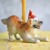 A Golden Retriever Cake Topper wearing a party hat, supporting a yellow candle with its tail, set against a blurred background with scattered confetti.