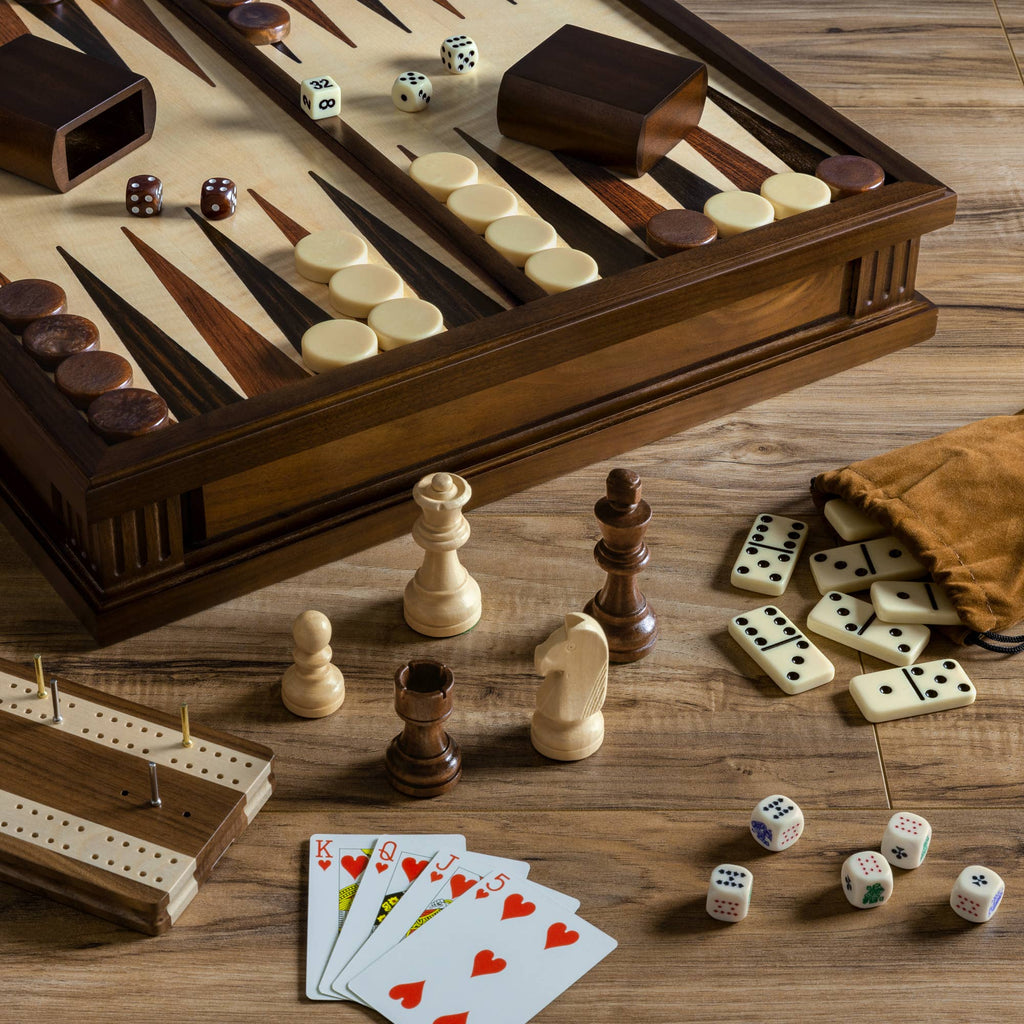 A wooden table displays assorted board game pieces, including the Heirloom Edition: 7-in-1 set (Chess, Checkers, Backgammon, Cribbage, Poker Dice, Cards, Dominoes), with all items arranged neatly in a walnut cabinet and brown pouch.