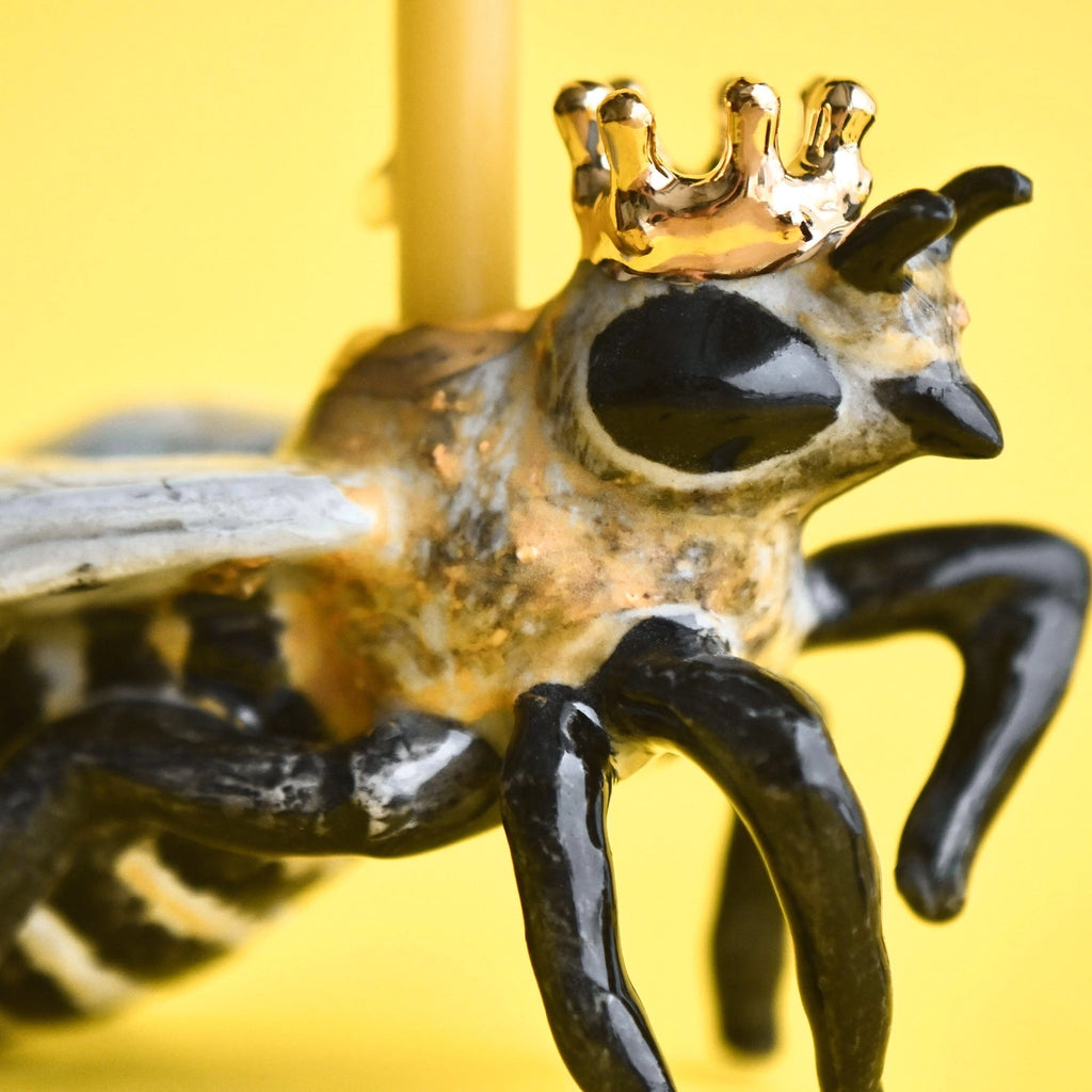 A close-up of the Queen Bee Cake Topper, a whimsical, hand-painted sculpture depicting a frog wearing a small golden crown, against a bright yellow background.