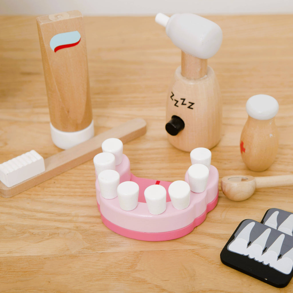 The Wooden Dentist Set is a pretend play toy featuring a wooden teeth model, toothbrush, toothpaste, dental tools, and a tooth x-ray, all displayed on a wooden surface.