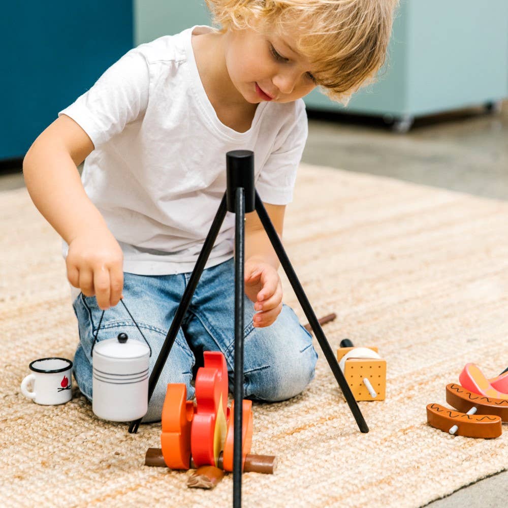 A young child enjoys indoor camping on a beige rug with the Wooden Camp Fire Set, featuring toy logs, flames, marshmallows, and a small kettle—perfect for immersive pretend play.