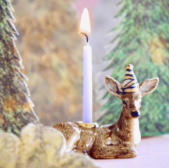 A fine porcelain deer cake topper with a lit white taper candle on its back, set against a soft-focus background of pine trees.