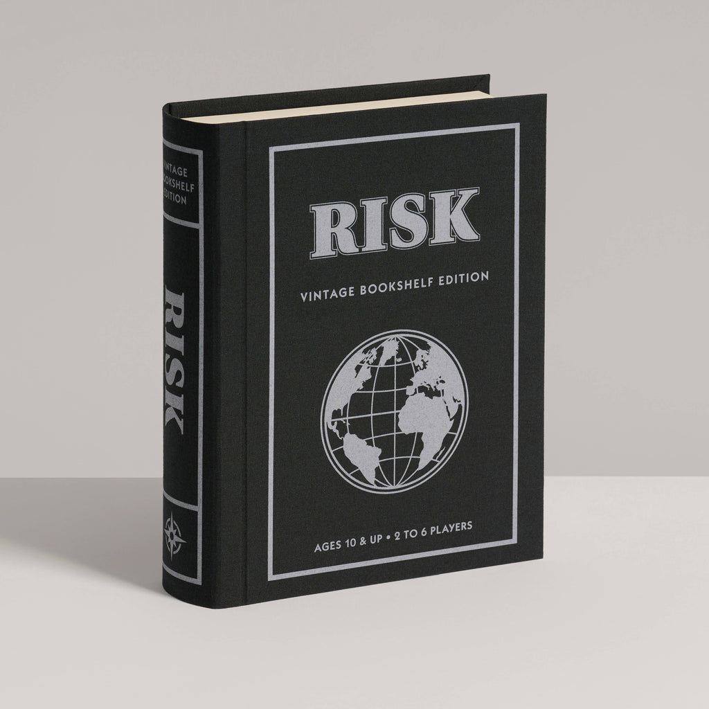 A black, book-shaped box labeled "Board Game: Risk Vintage Bookshelf Edition" features a white globe and classic RISK cards, designed to resemble a vintage hardcover book for stylish bookshelf display.